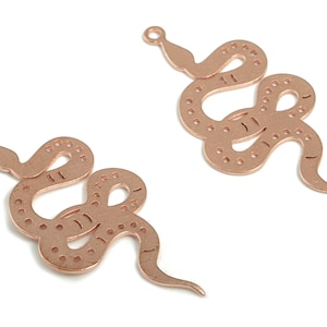 Puede incluir: Dos colgantes de serpiente de color cobre. Cada colgante tiene forma de serpiente estilizada con un lazo en la parte superior para colgar. Las serpientes tienen una serie de pequeños agujeros espaciados en sus cuerpos.