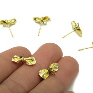 May include: Gold-colored bow-shaped stud earrings. The earrings have a post for insertion into the ear. The earrings are small and delicate, with a polished finish. The earrings are shown on a white background.
