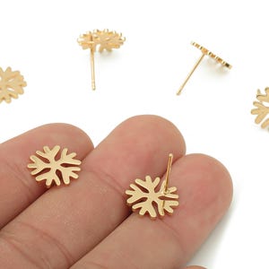 May include: Gold-colored snowflake stud earrings. The earrings are displayed on a hand against a white background. The earrings have a post and are made of a shiny metal. The snowflake design is intricate and detailed.