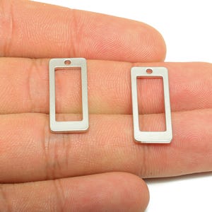 Puede incluir: Dos colgantes rectangulares plateados con forma de smartphone. Cada colgante tiene un marco rectangular con un pequeño agujero en la parte superior. Los colgantes se muestran en una mano.