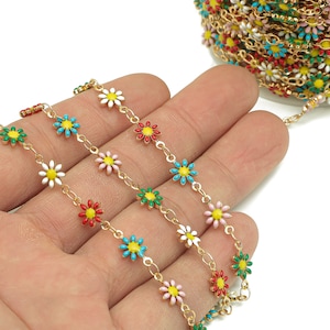 Könnte beinhalten: Eine goldene Kette mit bunten Email-Gänseblümchen. Die Kette wird auf einer Hand gezeigt, wobei die Blumen zum Betrachter zeigen.