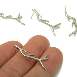 May include: Silver-toned, abstract stud earrings in a branch-like design. The earrings have a post backing and are displayed on a white background. One earring is held in a hand, showcasing the jewelry's shape and detail.