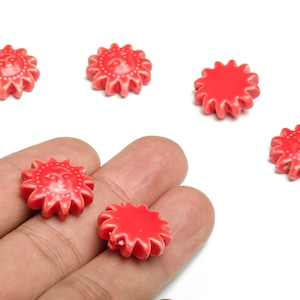 Peut inclure: Perles rouges en forme de soleil avec un visage. Les perles ont un bord texturé et strié. Certaines perles sont dans la main d'une personne. Les perles mesurent moins de 2,5 cm de diamètre.