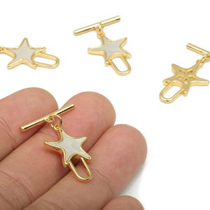 May include: Gold-colored star-shaped charms with a white center and a small clasp. The charms are attached to a gold bar. The image shows several charms, with one held in a hand.