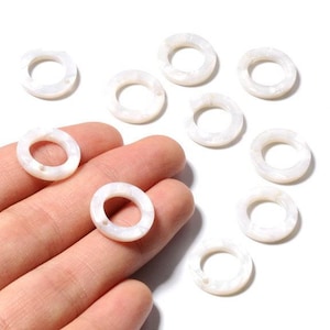 Könnte beinhalten: Eine Sammlung weißer, ringförmiger Perlen mit schimmerndem Glanz. Die Perlen liegen auf einer weißen Oberfläche, einige werden in einer Hand gehalten. Die Perlen haben ein Loch in der Mitte und werden wahrscheinlich für Schmuckherstellung oder Basteln verwendet.