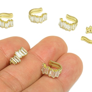 Può includere: Polisini per orecchie color oro con pietre di zirconia cubica trasparente taglio baguette. I polsini per orecchie sono mostrati in diverse posizioni, alcuni su una mano e altri su sfondo bianco. I gioielli hanno un design moderno e minimalista.