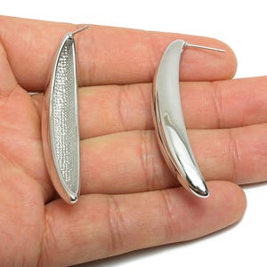 May include: A pair of silver-coloured, curved earrings. One earring has a textured interior, while the other is smooth. The earrings are held in a person's hand against a white background. The earrings have a post for pierced ears.
