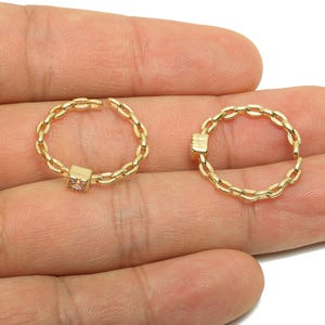 May include: Two gold-coloured chain rings with a square accent. One ring is open, and the other is closed. The rings are held in a hand against a white background. The square accent appears to be a light pink colour.