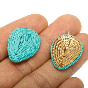 May include: Two turquoise raffia teardrop-shaped earrings. One earring is a solid teardrop shape, while the other has a gold-coloured metal backing with a post for piercing. The earrings are held in a hand.