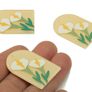 Puede incluir: Tres dijes acrílicos de color amarillo claro con un diseño floral. Cada dije presenta un lirio de agua blanco con estambres amarillos y hojas verdes. Los dijes tienen forma de rectángulo redondeado.