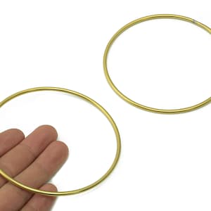 May include: Two gold-coloured metal hoops, one held by a hand, are displayed against a white background. The hoops are circular and appear to be made of a thin, shiny material. The hoops are likely used for crafting or jewellery making.