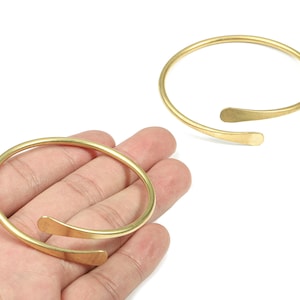 Könnte beinhalten: Zwei goldfarbene, offene Armreifen. Jedes Armband hat ein einfaches, abgerundetes Band mit abgeflachten, gebogenen Enden. Die Armbänder werden auf weißem Hintergrund präsentiert.