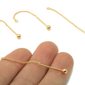 Puede incluir: Componentes de joyería dorados. Los componentes incluyen una pequeña bola al final de una barra recta y una cadena. Las joyas se muestran sobre un fondo blanco. Una pieza está sujeta por los dedos de una persona.