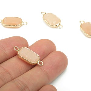May include: Four peach-colored, rectangular gemstone connectors with gold-colored metal edging and loops. One connector is held in a hand. The gemstones have a textured surface. Ideal for jewelry making.