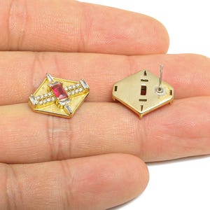 May include: Two gold-colored stud earrings. Each earring has a square shape with a central red gemstone and smaller white gemstones. One earring is face up, the other is face down, showing the post.
