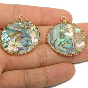 Könnte beinhalten: Zwei runde Abalone-Muschel-Anhänger mit goldfarbenem Rand. Die schillernden Muscheln zeigen eine Mischung aus Blau, Grün und Lila. Jeder Anhänger hat oben eine kleine Schlaufe zum Aufhängen, ideal für Schmuck.