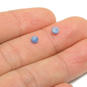 Puede incluir: Dos pequeñas piedras preciosas redondas, de color azul claro, se muestran en los dedos de una persona. Las piedras tienen un aspecto ligeramente texturizado. Podrían usarse para la fabricación de joyas.