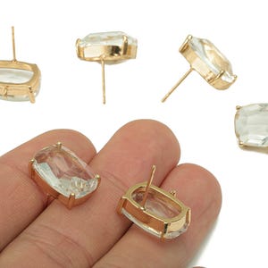 May include: Clear, rectangular-cut gemstone stud earrings with gold-colored settings. The earrings are displayed on a white background, showcasing their facets and the gold metal. The studs are a classic jewelry item.