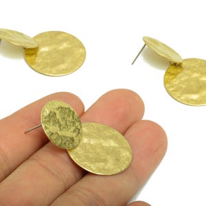 Peut inclure: Boucles d'oreilles en métal doré martelé. Chaque boucle d'oreille comporte deux formes circulaires reliées. Les boucles d'oreilles sont présentées sur fond blanc. Les boucles d'oreilles sont tenues dans une main.