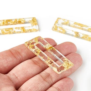 May include: Rectangular clear resin jewelry components with gold leaf flakes. The components have a rectangular cutout in the center. Three pieces are visible, with one held in a hand against a white background.