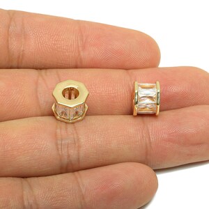 以下が含まれることがあります： クリアな長方形カットの石が付いた、2つの金色のジュエリービーズ。1つのビーズは八角形で、もう1つは円筒形です。ビーズは白い背景に手のひらに表示されています。