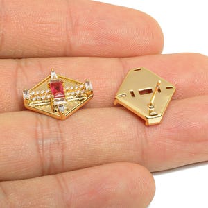 May include: Gold-toned stud earrings with a geometric design. Each earring features a central rectangular red gemstone, surrounded by smaller white gemstones and a diamond-shaped frame. One earring is shown with the post exposed.