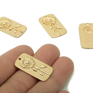 May include: Gold-colored rectangular pendants with a sunflower design. The pendants have a detailed embossed sunflower with leaves and stem. One pendant is held in a hand, and the others are scattered on a white surface.