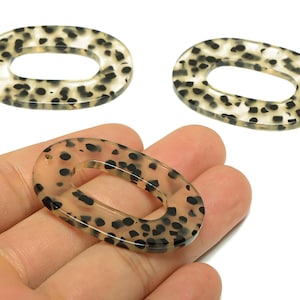 May include: Three oval-shaped beads with a translucent beige base and black spotted pattern. One bead is held in a hand. These beads are suitable for jewelry making and crafting projects.