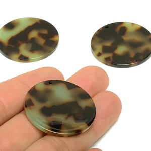 Pode incluir: Três contas redondas e planas com um padrão de tartaruga em tons de castanho, bege e verde. Uma conta é segurada na mão, e todas têm um pequeno orifício para enfiar. Estas contas são adequadas para fazer joias.