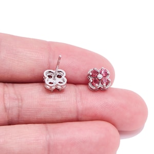 Può includere: Un paio di orecchini a lobo a forma di fiore. Ogni orecchino presenta un gruppo di pietre preziose rosa circondate da un alone di pietre più piccole e trasparenti. Gli orecchini sono incastonati in un metallo color argento e sono esposti su sfondo bianco.