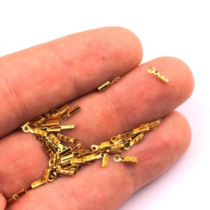 Puede incluir: Una colección de pequeños conectores eléctricos dorados. Los conectores son rectangulares con un pequeño bucle en un extremo. Se sostienen en la mano de una persona sobre un fondo blanco.