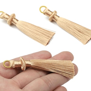 May include: Three gold-colored tassels with a looped top and a knotted detail. The tassels are made of fine, silky threads, and one is held in a hand. These decorative tassels are suitable for jewelry making or craft projects.