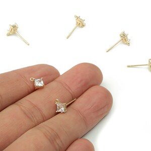 May include: Gold-colored stud earrings with a square, clear gemstone. Several earrings are scattered on a white surface, with two held in a hand. The earrings have a post and a small, decorative element.
