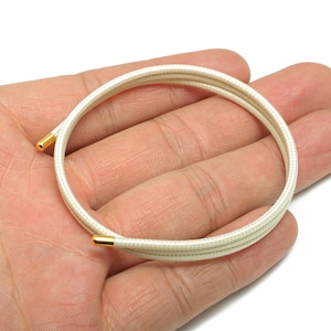 May include: A cream-colored bracelet with gold-colored metal ends. The bracelet is made of a woven, cord-like material and is circular in shape. The bracelet is held in a person's hand.
