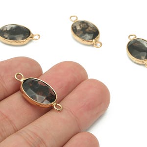 May include: Oval-shaped gemstone connector beads with a gold-colored bezel. The gemstones have a dark, mottled appearance with shades of black, brown, and gray. Each bead has a loop on either end for stringing.