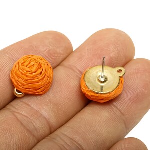 May include: A pair of orange woven stud earrings. Each earring has a round, woven orange design with a gold-coloured metal backing and post. The earrings are shown against a neutral background.
