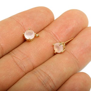 May include: A pair of gold-colored stud earrings, each featuring a round, pale pink gemstone. The earrings are set in a four-prong setting, with a post and clasp for secure wear. The earrings are displayed on a light skin tone.