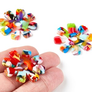 Könnte beinhalten: Drei bunte blütenförmige Perlen mit einem Loch in der Mitte. Die Perlen sind eine Mischung aus Rot, Blau, Gelb, Grün und Weiß. Eine Perle wird in einer Hand gehalten.