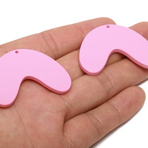 Puede incluir: Dos colgantes rosas pálidos en forma de bumerán con pequeños agujeros para hacer joyas. Los colgantes son lisos y curvos. Se sostienen en la mano de una persona.