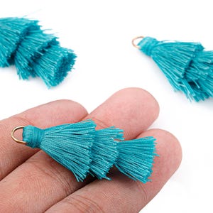 May include: Three turquoise tiered tassels with gold-colored loops for jewelry making or crafting. One tassel is held in a hand, while two others are displayed separately against a white background. The tassels are made of fine, silky threads.