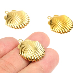 May include: Three gold-colored seashell charms with a textured, ridged design. One charm is held in a hand, while the other two are on a white background. Each charm has a small loop at the top for attaching to jewelry.