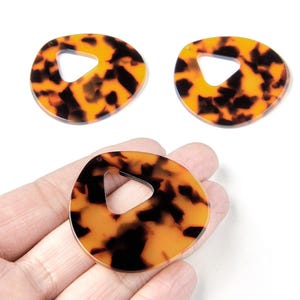 Peut inclure: Trois pendentifs en acrylique à motif écaille de tortue. Chaque pendentif a une forme organique avec une découpe triangulaire. Les pendentifs sont bruns et ambrés, avec une finition brillante. Idéal pour la création de bijoux.