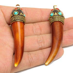 May include: Two brown pendants with a horn-like shape. Each pendant has a decorative cap with turquoise and red accents, and a gold-colored bail. The pendants are held in a person's hand.