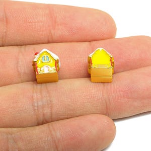 以下が含まれることがあります： 2つのミニチュアハウスチャーム。家は黄色で、白い屋根、赤いアクセント、金のトリムが付いています。1つのチャームは正面から、もう1つは背面から見たものです。チャームは手で持たれています。