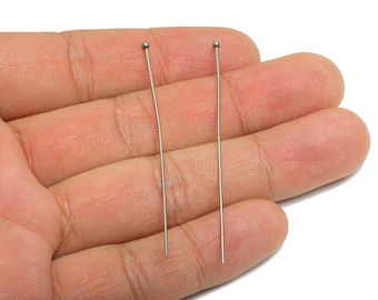 May include: Two silver-colored head pins with a small ball at the top. The pins are held in a person's hand, with the pins' thin, straight shafts extending downwards. The background is a plain white surface.