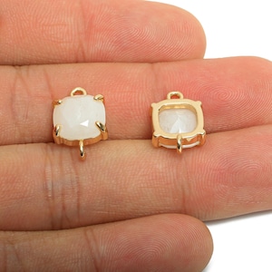 May include: Two square-shaped jewelry pendants with a gold-colored frame and a white, faceted stone. Each pendant has a small loop at the top and a small loop at the bottom. The pendants are displayed on a light-colored background.