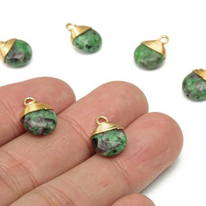 May include: Green and red gemstone pendants with gold-colored caps and loops. The pendants are small, round, and have a natural, speckled appearance. Several pendants are scattered on a white background, with some held in a hand.