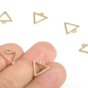 Puede incluir: Varios pequeños dijes triangulares dorados con un lazo en la parte superior para hacer joyas. Algunos dijes se sostienen en una mano, mientras que otros están esparcidos sobre una superficie blanca. Los dijes son geométricos y minimalistas.