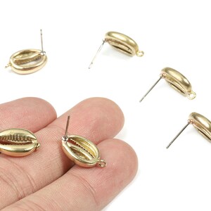 May include: Gold-colored seashell stud earrings. The earrings are shaped like small seashells with a detailed texture. The posts are silver-colored. Several earrings are scattered on a white background, with two held in a hand.