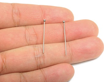 May include: Two silver-colored head pins with a small ball at the top. The pins are held in a person's hand, with the pins' straight shafts extending downwards. The background is a light, neutral color.